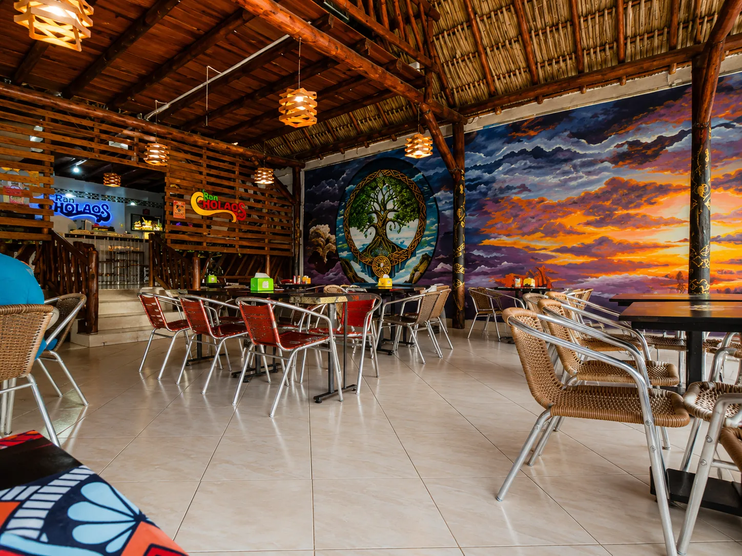 Interior del local de RanCholaos en Villavicencio: techo de madera y palma, los murales del árbol de la vida y el atardecer, mesas con sillas y los letreros de la marca al fondo