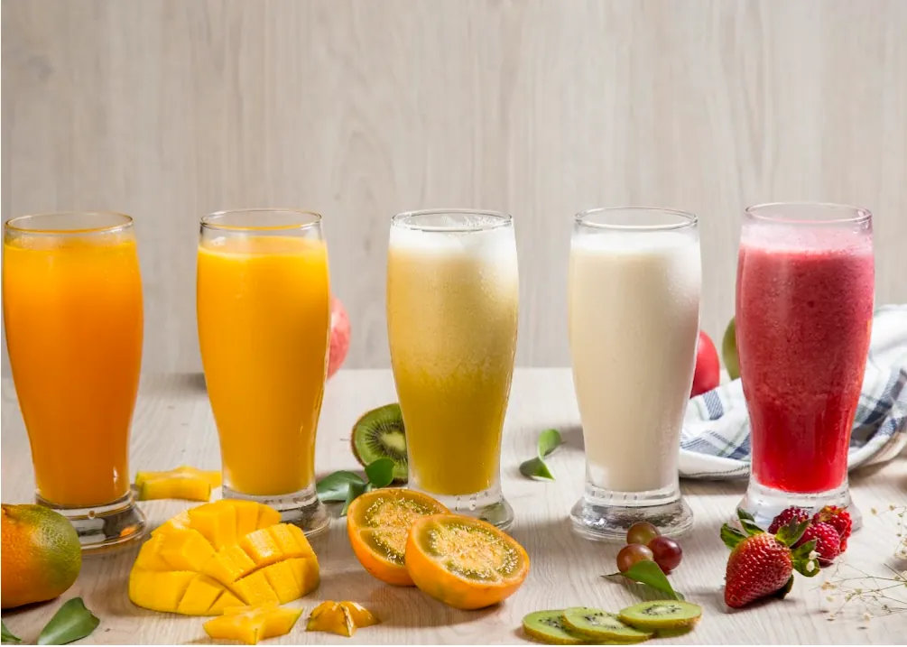 Hilera de jugos naturales de fruta fresca —mango, naranja, mora y más— en vasos altos